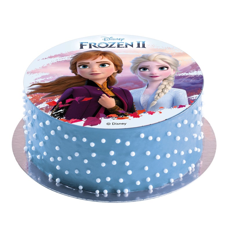 Pode incluir: Um bolo azul e branco com uma imagem comest&iacute;vel redonda de Elsa e Anna de Frozen II da Disney. A imagem tem um fundo de p&ocirc;r do sol rosa e roxo com montanhas ao longe. O texto "Disney FROZEN II" est&aacute; impresso no bolo.