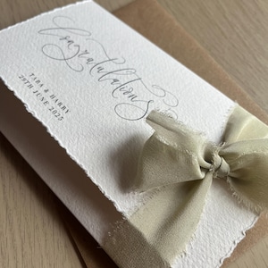 May include: A white card with the word "Congratulations" in elegant script, tied with a light green ribbon. The card includes the names "Tara & Harry" and the date "29th June 2025". The card is on a brown envelope.