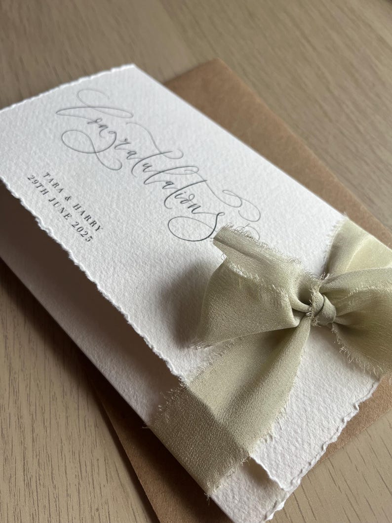 May include: A white card with the word "Congratulations" in elegant script, tied with a light green ribbon. The card includes the names "Tara & Harry" and the date "29th June 2025". The card is on a brown envelope.
