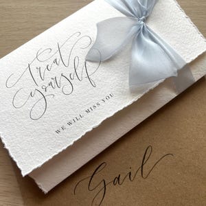 May include: A white card with the words "Treat Yourself" and "We will miss you" in elegant script, tied with a light blue ribbon. A brown envelope with the name "Gail" in matching script is underneath.