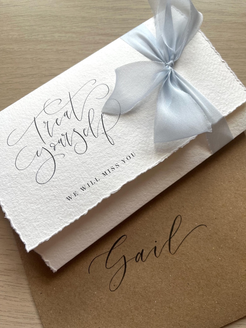 May include: A white card with the words "Treat Yourself" and "We will miss you" in elegant script, tied with a light blue ribbon. A brown envelope with the name "Gail" in matching script is underneath.