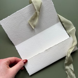 May include: A handmade, textured white paper card with a torn edge, held open to reveal the inside. A light green silk ribbon is tied around the card. The background is a solid, muted green.