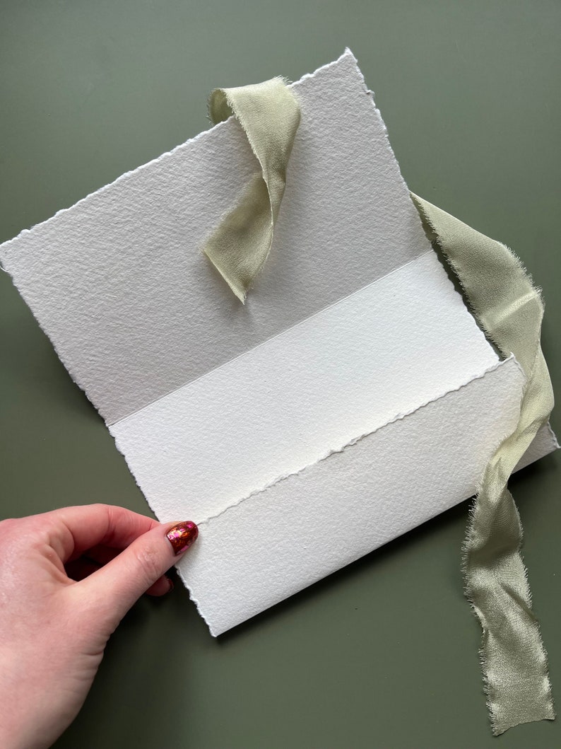 May include: A handmade, textured white paper card with a torn edge, held open to reveal the inside. A light green silk ribbon is tied around the card. The background is a solid, muted green.