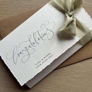 May include: A white card with the word "Congratulations" in elegant script, tied with a light green ribbon. The card includes the names "TARA & HARRY" and the date "29TH JUNE 2025". The card is placed on a brown envelope.