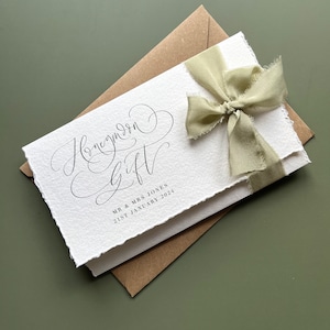 May include: A white card with the words "Honeymoon Gift" in elegant script, tied with a light green ribbon. The card is layered over a brown envelope, with the names "MR & MRS JONES" and the date "21ST JANUARY 2024" printed below.