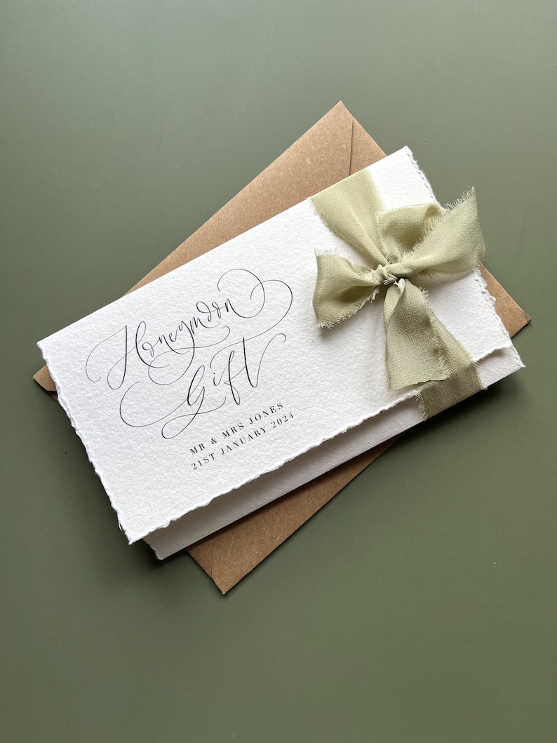 May include: A white card with the words "Honeymoon Gift" in elegant script, tied with a light green ribbon. The card is layered over a brown envelope, with the names "MR & MRS JONES" and the date "21ST JANUARY 2024" printed below.