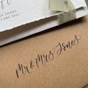 May include: Close-up of wedding stationery. A white card with torn edges is tied with a sage green ribbon. The card has elegant calligraphy. Below, a brown envelope is inscribed with "Mr & Mrs Jones" in black script.