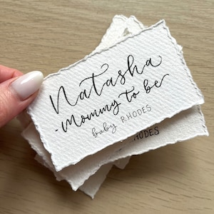 Könnte beinhalten: Ein Stapel weißer Papierkarten mit dem Text "Natasha - Mommy to be" und "baby RHODES" in schwarzer Tinte.