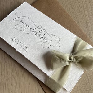 Könnte beinhalten: Eine weiße Karte mit dem Wort "Glückwunsch" in eleganter Schrift, kombiniert mit einem braunen Umschlag und einem salbeigrünen Band. Die Karte enthält die Namen "Tara & Harry" und das Datum "29. Juni 2025".