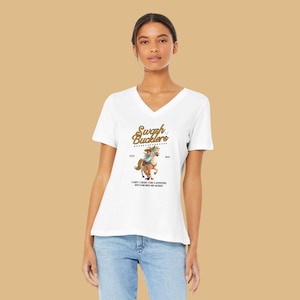 May include: White V-neck t-shirt with a cartoon illustration of a person riding a horse. The shirt displays the text "Swash Bucklers" in gold script above the image, with additional text below. The shirt is worn by a person.