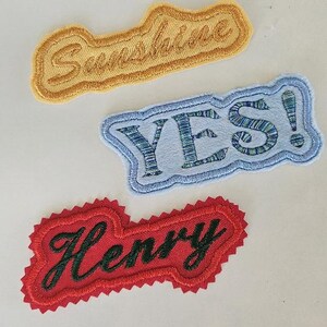 Könnte beinhalten: Drei bestickte Stoffaufnäher. Der obere Aufnäher ist gelb mit dem Wort Sunshine in Schreibschrift. Der mittlere Aufnäher ist hellblau mit dem Wort YES! in Dunkelblau. Der untere Aufnäher ist rot mit dem Namen Henry in dunkelgrüner Schreibschrift.