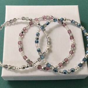 May include: Three delicate bracelets made with silver and iridescent beads in shades of blue, pink, and grey.