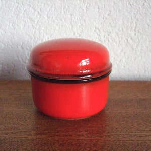 Könnte beinhalten: Eine rote Keramikdose mit schwarzem Rand und Deckel. Die Dose ist rund und hat eine glatte, glänzende Oberfläche.