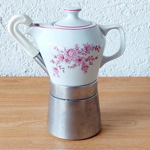 Könnte beinhalten: Eine weiß-silberne Espressomaschine mit floralem Design. Der obere Teil ist weiß mit rosa Blumenmustern und rotem Rand. Der Griff ist weiß mit geschwungenem Design. Der Boden ist silberfarben.
