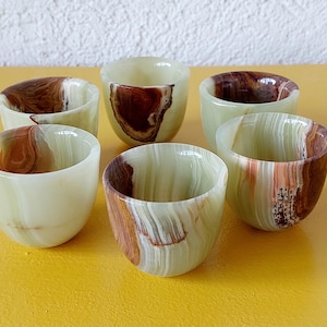 Può includere: Sei piccole tazze rotonde in onice con un colore di base verde chiaro e venature marroni e bianche. Le tazze sono disposte su una superficie gialla. Le tazze hanno una finitura liscia e lucida.