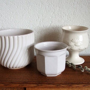 K&ouml;nnte beinhalten: Drei wei&szlig;e Keramik-Blument&ouml;pfe, zwei mit glatten Oberfl&auml;chen und einer mit einem wellenf&ouml;rmigen Design. Die T&ouml;pfe stehen auf einer Holzoberfl&auml;che.