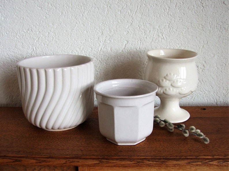 K&ouml;nnte beinhalten: Drei wei&szlig;e Keramik-Blument&ouml;pfe, zwei mit glatten Oberfl&auml;chen und einer mit einem wellenf&ouml;rmigen Design. Die T&ouml;pfe stehen auf einer Holzoberfl&auml;che.