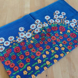 Könnte beinhalten: Gefaltetes blaues Textil mit Blumenmuster. Das Design zeigt weiße Gänseblümchen, rote und orangefarbene Blumen und gelbe tulpenartige Formen. Die Blumen sind in Reihen auf blauem Hintergrund angeordnet.