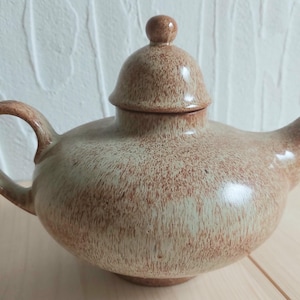 Può includere: Una teiera in ceramica marrone e beige con un coperchio. La teiera ha una glassa macchiata e un corpo arrotondato.