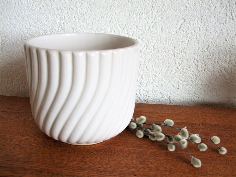 K&ouml;nnte beinhalten: Wei&szlig;er Keramiktopf mit wellenf&ouml;rmigem, geripptem Design. Der Topf steht auf einer Holzoberfl&auml;che mit einem Zweig Weidenk&auml;tzchen.