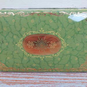 Può includere: Una scatola di metallo verde con un bordo dorato e un design decorativo che presenta un ovale dorato con tre fiori dorati. La scatola ha uno sfondo verde marmorizzato con piccoli fiori bianchi.