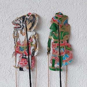 Könnte beinhalten: Zwei traditionelle indonesische Schattenspielfiguren. Die Puppe links ist weiß, schwarz und goldfarben, mit roten und goldenen Akzenten. Die Puppe rechts ist grün, rot und goldfarben, mit blauen Akzenten. Beide Puppen sind auf Stäben montiert.