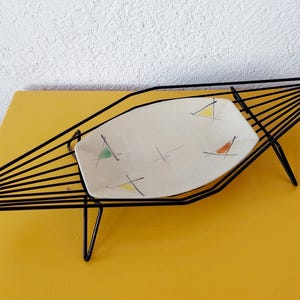 Könnte beinhalten: Ein Retro-Serviertablett mit einem schwarzen Metallrahmen und einer cremefarbenen Keramikschale. Die Schale hat abstrakte geometrische Designs in Gelb, Grün und Orange. Das Tablett steht auf einer gelben Oberfläche.