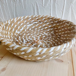 Könnte beinhalten: Ein runder, flacher Korb aus geflochtenem Naturmaterial mit weißen Streifen. Der Korb hat einen Durchmesser von etwa 30 cm.