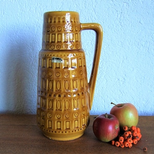 Puede incluir: Un jarrón de cerámica marrón con un patrón geométrico y un asa. El jarrón está sobre una mesa de madera con dos manzanas rojas y un racimo de bayas naranjas.