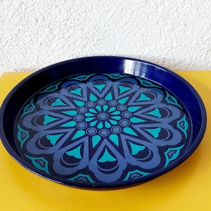 Pode incluir: Uma bandeja redonda azul marinho com um padrão floral turquesa e azul. A bandeja está colocada sobre uma superfície amarela, com uma parede branca ao fundo. O design da bandeja apresenta um motivo central em forma de flor.