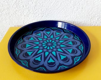 May include: A round, navy blue serving tray with a teal and blue floral pattern. The tray sits on a yellow surface, with a white wall in the background. The tray's design features a central flower-like motif.