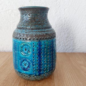 Op de afbeelding: Een getextureerde keramische vaas in turquoise en bruin. De vaas heeft een cilindrische vorm met een brede basis en een smallere hals. Het turquoise gedeelte heeft een patroon van cirkels en verticale lijnen.