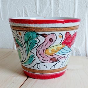 May include: A ceramic flower pot with a hand-painted design. The pot has a red rim and base, with a white body decorated with a colourful bird and floral motif. The bird has pink, yellow, blue, and red details. The pot is approximately 13 cm tall.