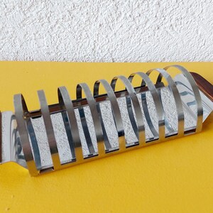 Può includere: Un portapane vintage in cromo e legno. Il portapane ha una base rettangolare con divisori metallici curvi. Le estremità del portapane sono in legno. È appoggiato su una superficie gialla e un muro bianco strutturato.