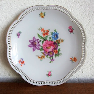 Puede incluir: Un plato de porcelana blanca con un borde festoneado y un delicado diseño floral. El plato presenta un ramo de flores rosas, moradas y amarillas con hojas verdes. El plato tiene un borde dorado y un fondo blanco.