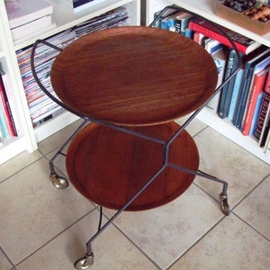 Puede incluir: Un carro de servicio de dos niveles con un marco de metal negro y dos bandejas redondas de madera. El carro tiene cuatro ruedas y está colocado frente a una estantería.