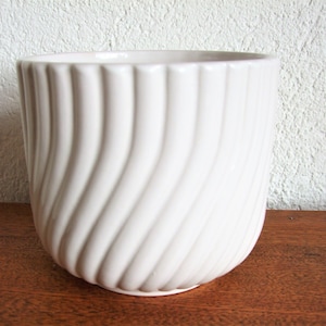 K&ouml;nnte beinhalten: Wei&szlig;er Keramiktopf mit wellenf&ouml;rmigem, geripptem Design. Der Topf ist leer und steht auf einer Holzoberfl&auml;che.