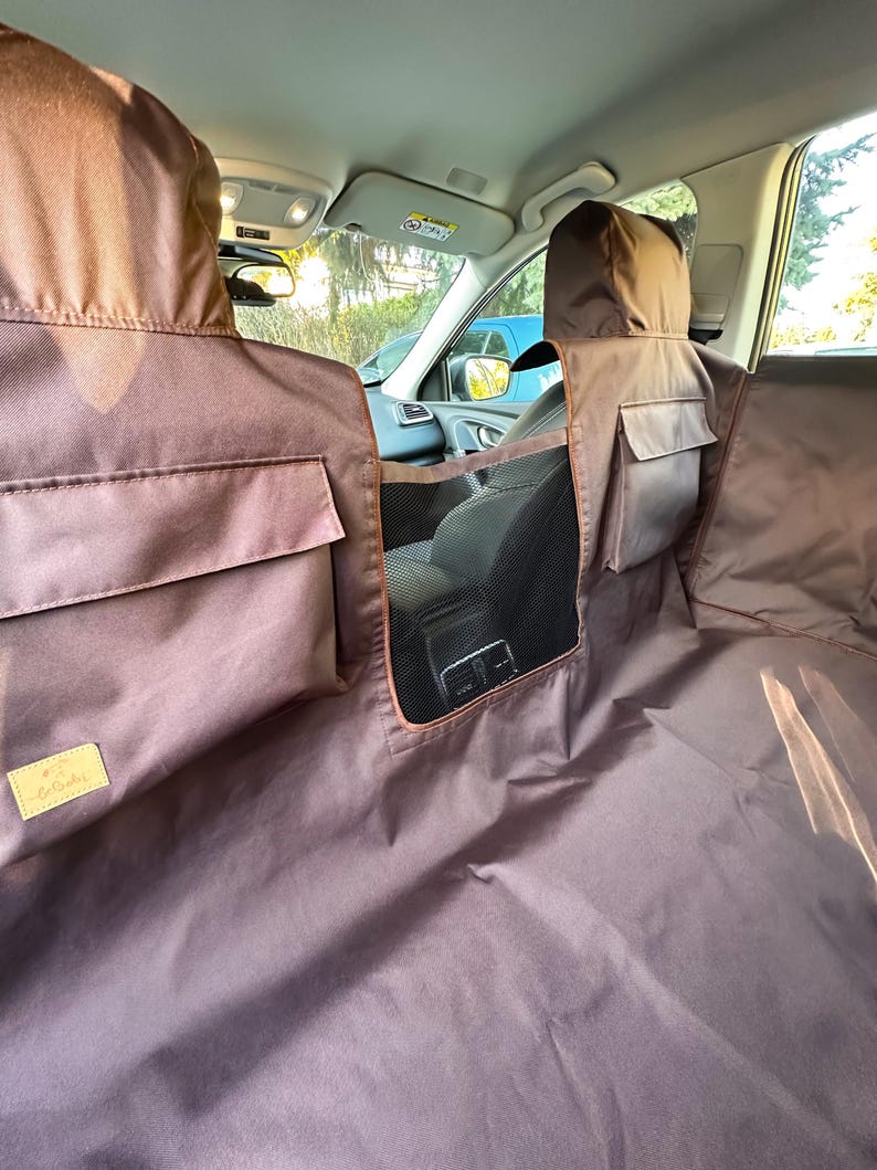May include: Brown car seat cover with a mesh divider and pockets. The cover is made of a water-resistant material and has a logo on the bottom left corner.