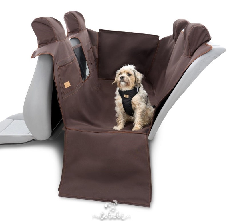 May include: Brown car seat protector with a small dog sitting on it. The protector has two headrest covers and a pocket on the back. The dog is wearing a black harness.