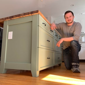 May include: A light green kitchen island with a butcher block countertop and six drawers. The island has black drawer pulls and sits on a wood floor.