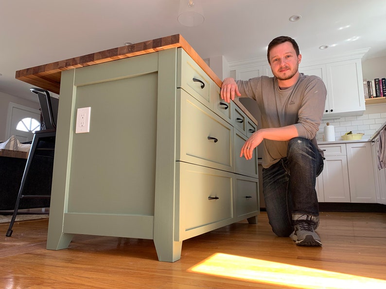 May include: A light green kitchen island with a butcher block countertop and six drawers. The island has black drawer pulls and sits on a wood floor.