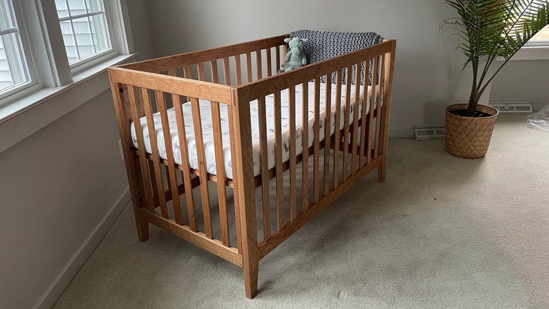 May include: A wooden crib with a white and gray patterned mattress and a stuffed animal. The crib is in a room with a window and a plant in a woven basket.
