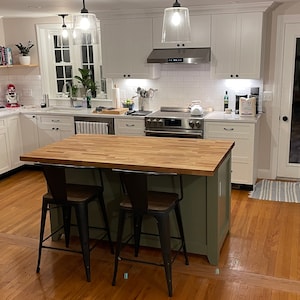 May include: A kitchen with white cabinets, stainless steel appliances, and a green island with a butcher block countertop. The island has two black metal bar stools. The kitchen has hardwood floors and a glass door leading to the backyard.