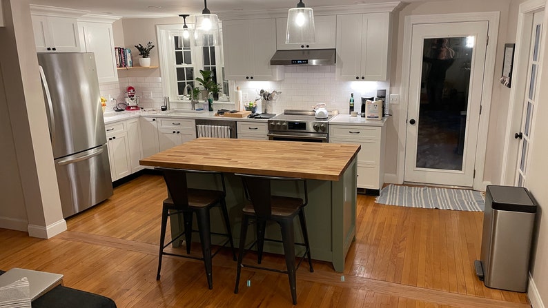 May include: A kitchen with white cabinets, stainless steel appliances, and a green island with a butcher block countertop. The island has two black metal bar stools. The kitchen has hardwood floors and a glass door leading to the backyard.