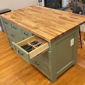 May include: A green kitchen island with a butcher block countertop and a drawer open revealing a knife block. The island has several drawers and a white electrical outlet.