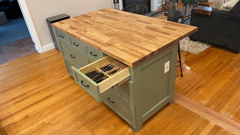 May include: A green kitchen island with a butcher block countertop and a drawer open revealing a knife block. The island has several drawers and a white electrical outlet.