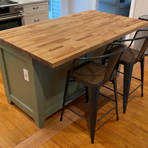 May include: A green kitchen island with a wood countertop and two black metal bar stools. The island has a white electrical outlet on the side.