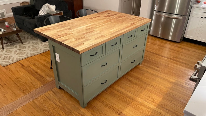 May include: A green kitchen island with a wood countertop and six drawers. The island has black drawer pulls and is standing on a hardwood floor.