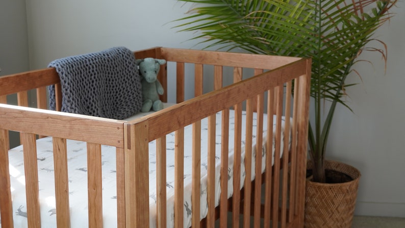 May include: A wooden crib with a white and gray patterned sheet and a gray knitted blanket. A small stuffed animal is sitting on the blanket.