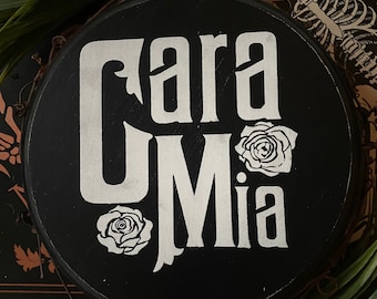 Puede incluir: Un letrero redondo de madera negro con las palabras "Cara Mia" escritas en letra cursiva blanca. El letrero está decorado con dos rosas blancas.